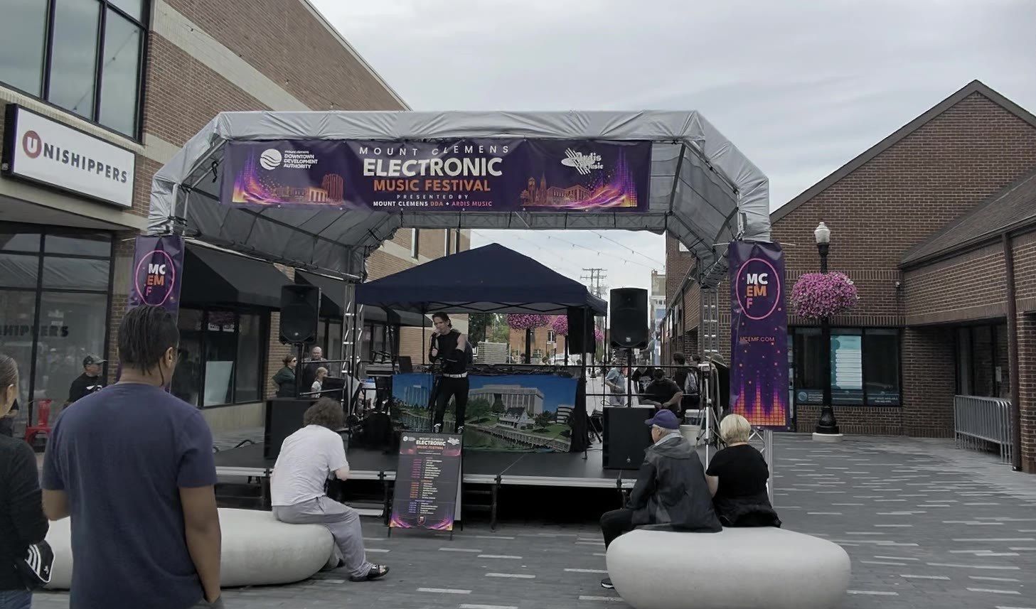 The MCEMF main stage and entry arch on Macomb Place, festivalgoers gathered on the plaza seating below the Mount Clemens Electronic Music Festival banner.