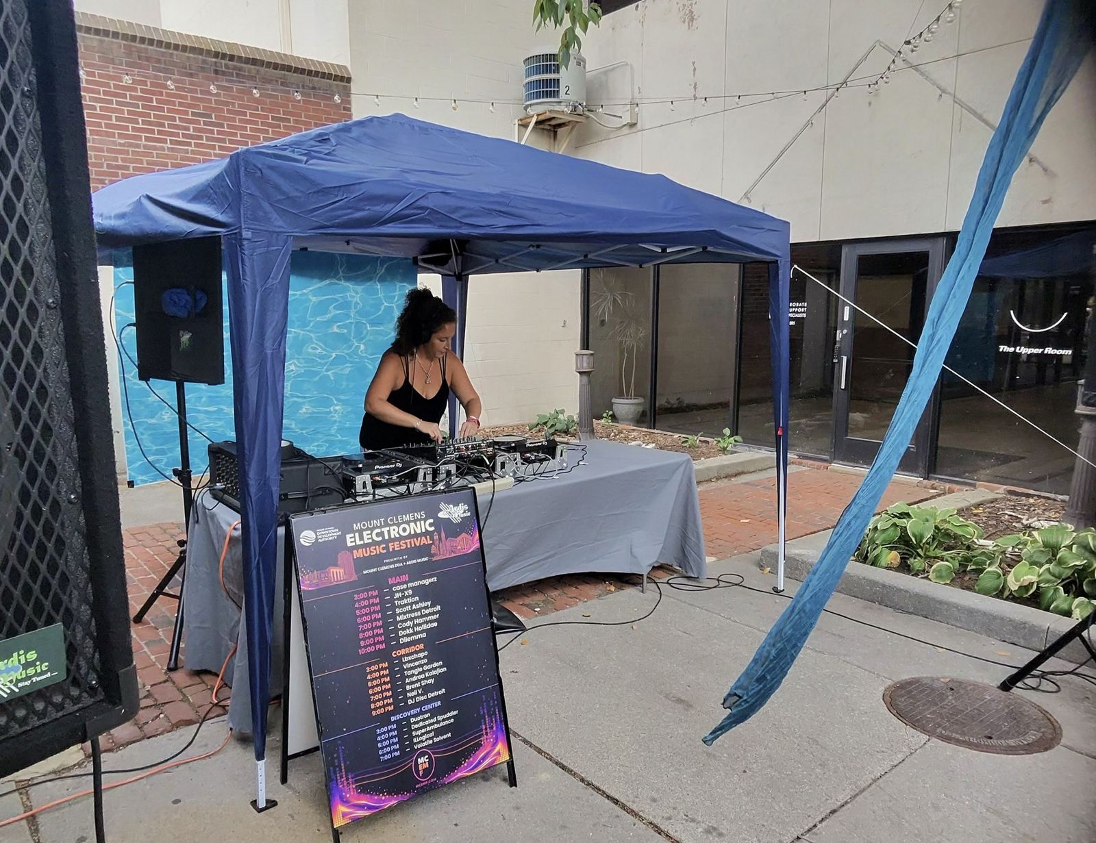 A DJ performing in the Corridor stage, set against the transformed passage's draped color and lighting, with the full festival set schedule posted alongside.