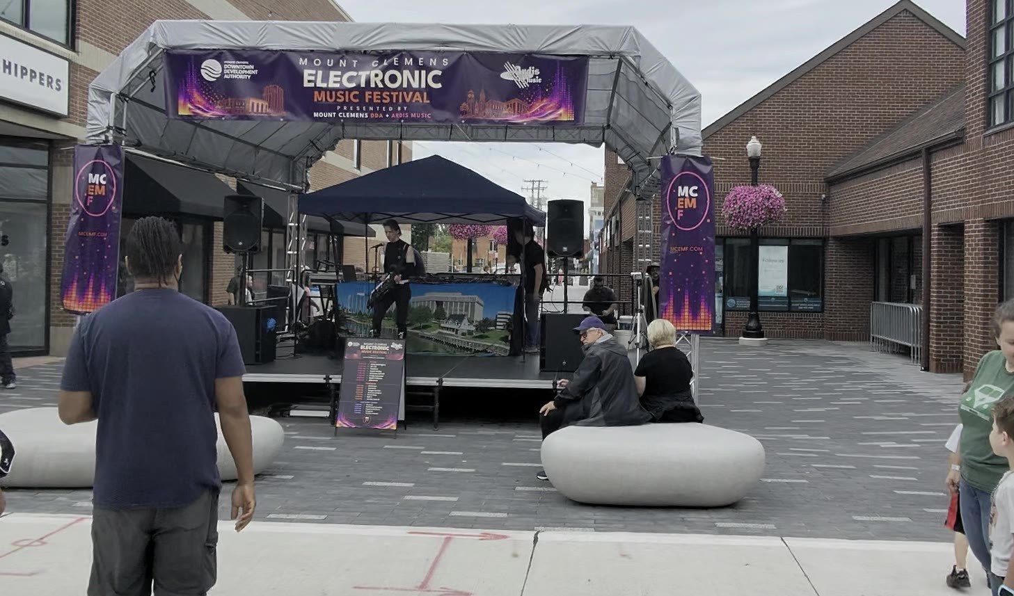 The MCEMF main stage and entry arch on Macomb Place in downtown Mount Clemens, with festivalgoers gathered on the plaza seating.